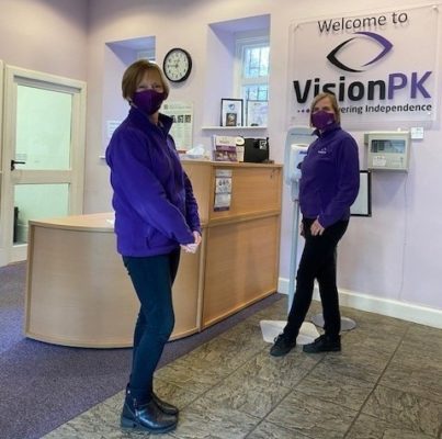 Two female VisionPK employees standing at VisionPK reception, looking at camera, wearing masks