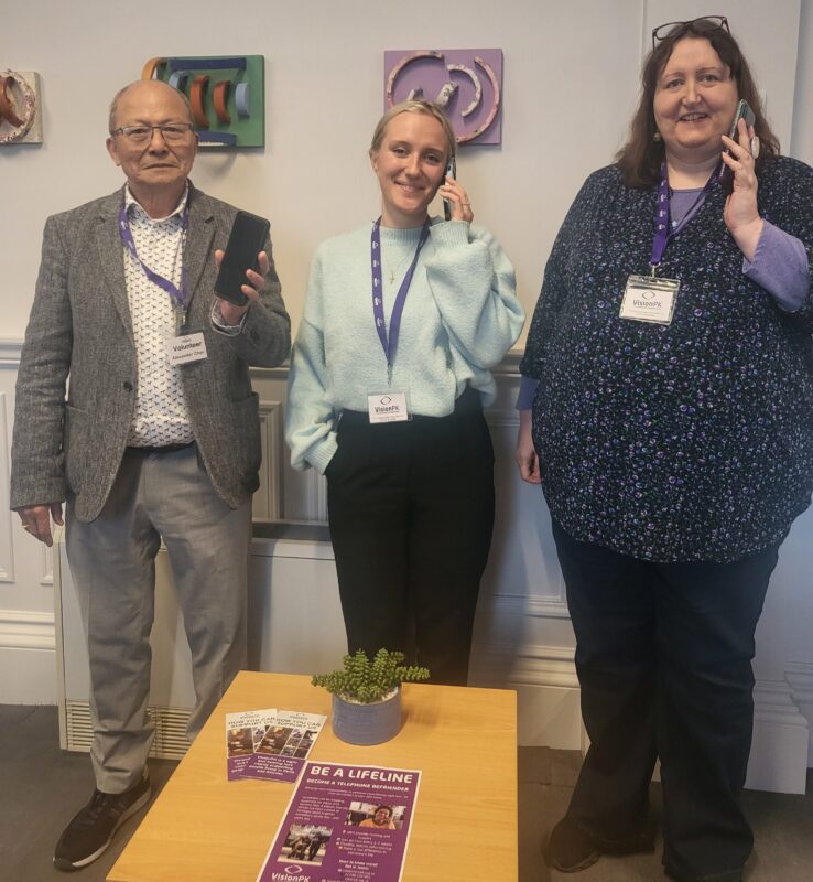 3 people standing facing the camera holding a mobile phone and smiling
