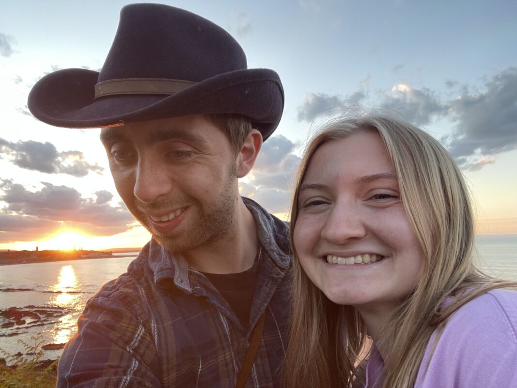 Man and woman smiling at the camera with coast and sunset in the background