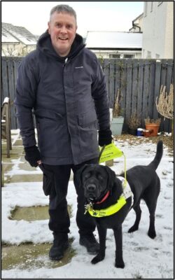 A man standing in the snow with his guide dog next to him.
