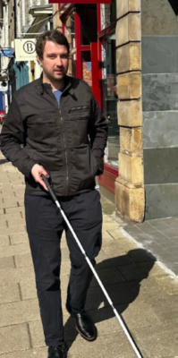 Man in his 30's with a visual impairment walking down a high street using a guide cane