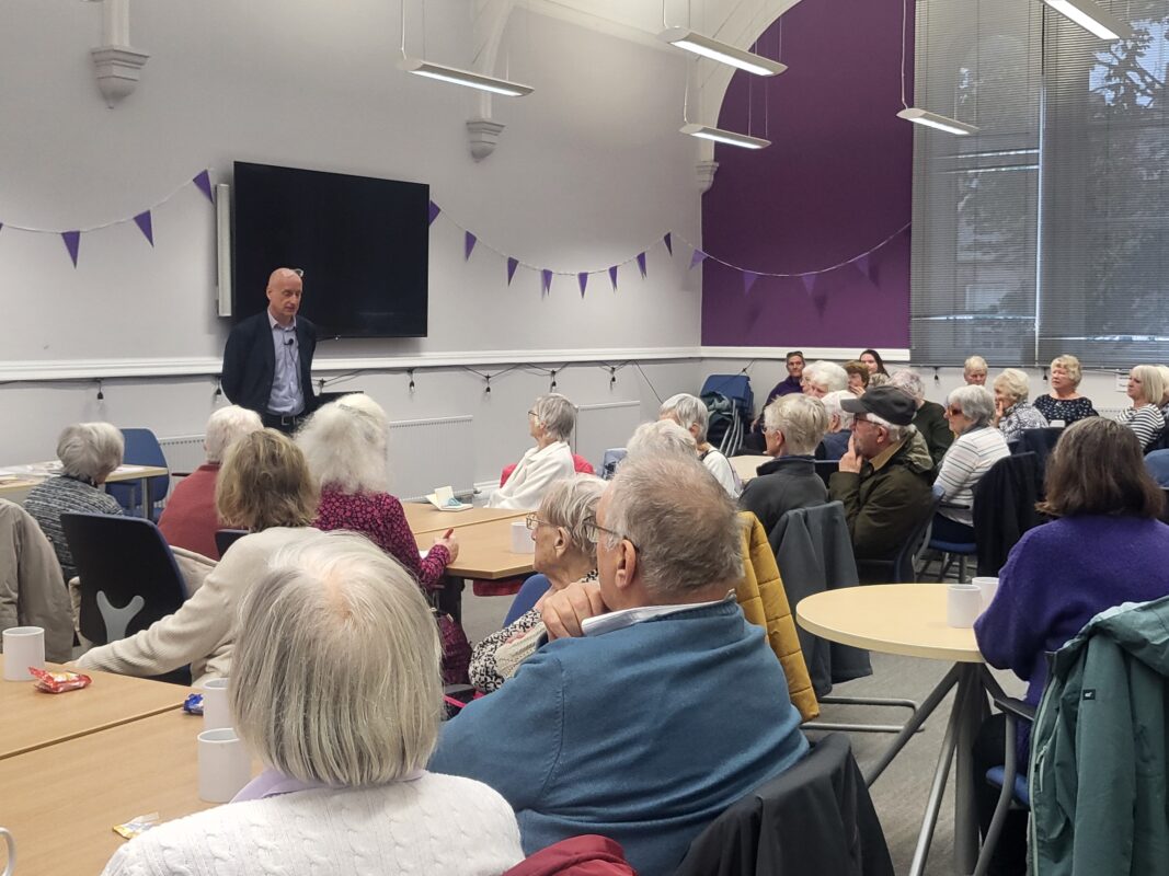 A large room with Dr Paul Johnstone standing at the fron hosting a talk to a large group of people all sitting and listening facing the speaker